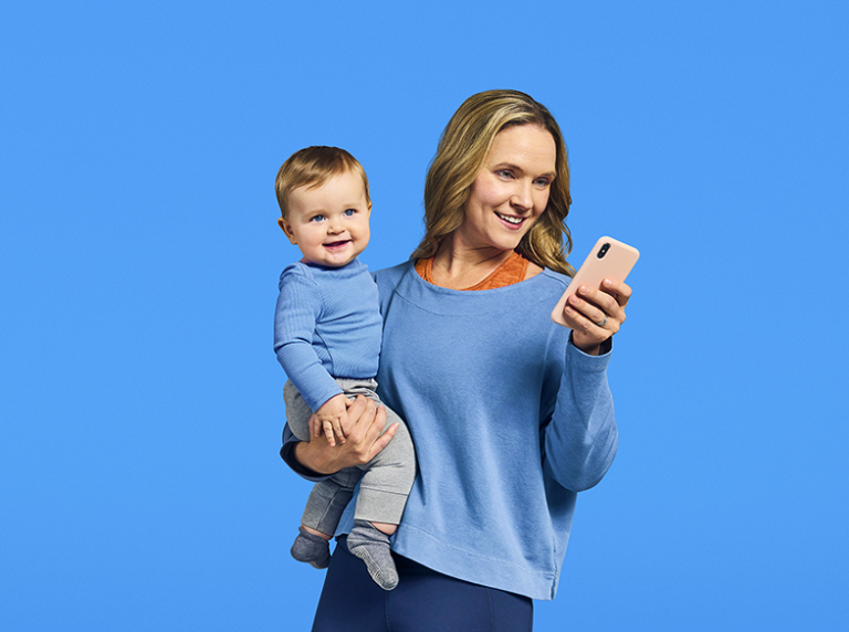 A woman holds a baby on one arm and holds a smartphone in her other hand.